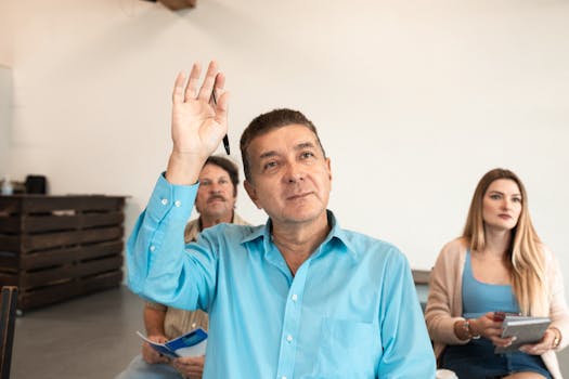 Adults learning in a classroom, with a man raising his hand to pose a question.
