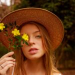 A woman wearing a straw hat holding yellow flowers in a serene outdoor setting.