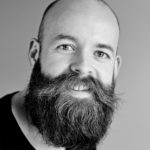 Black and white portrait of a smiling bald man with a large beard in a studio setting.